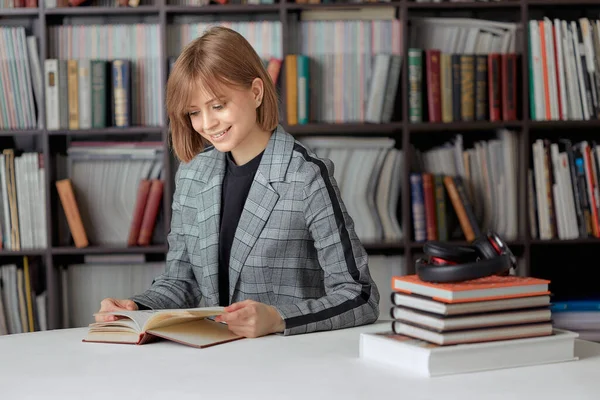 Frumoasă fată student lectură carte în bibliotecă cu stivă de cărți și căști wireless, pe fundalul raftului de bibliotecă — Fotografie, imagine de stoc