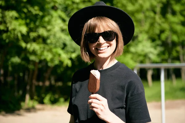 Pretty laughing young girl in sunglasses and hat eating ice cream in the park, photography for blog or ad