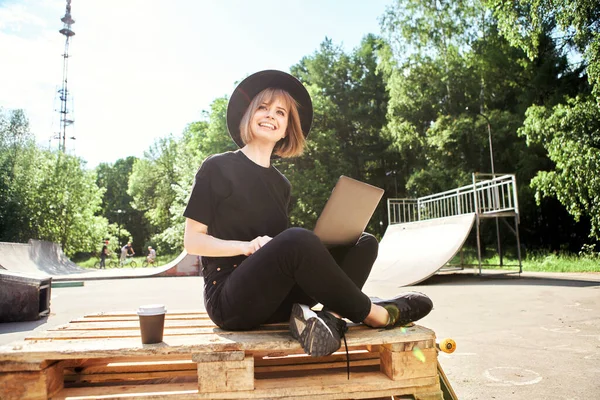 Creative young happy girl blogger work on laptop on content sitting on wooden pallet with cup and skateboard in park