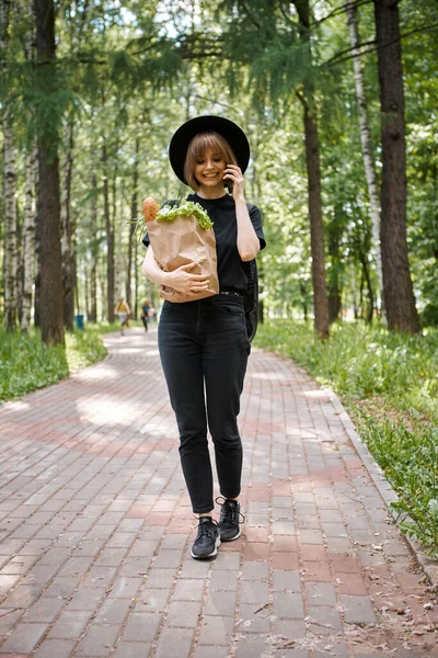 Young woman in hat with bag with vegetables communicate by phone walking in park, photography for blog or advertising