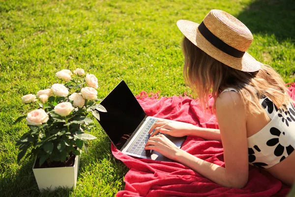 Creative young girl blogger in a hat working on a laptop on content on a green lawn with roses in summer