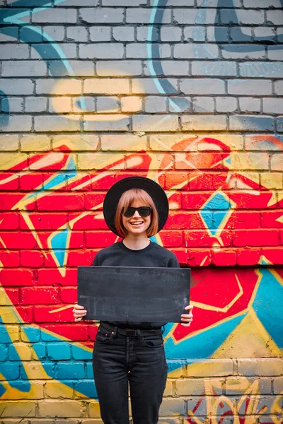 Muchacha alegre joven con cartel de cartón en el sombrero y gafas de sol de pie contra la pared de ladrillo con graffiti de colores — Foto de Stock