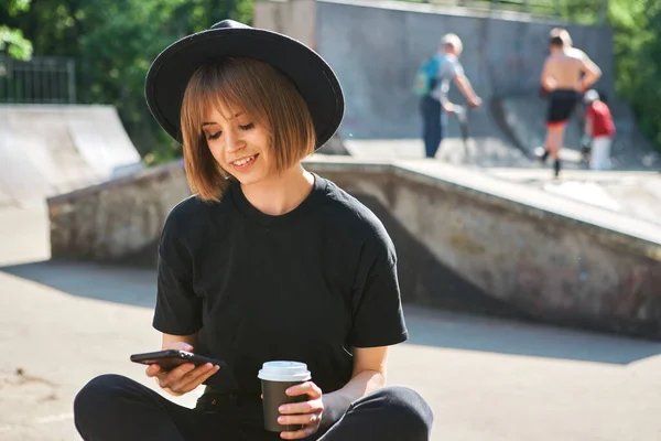 Pretty smiling woman blogger work on phone on content sitting with cup coffee to go in skatepark, photo for blog or ad
