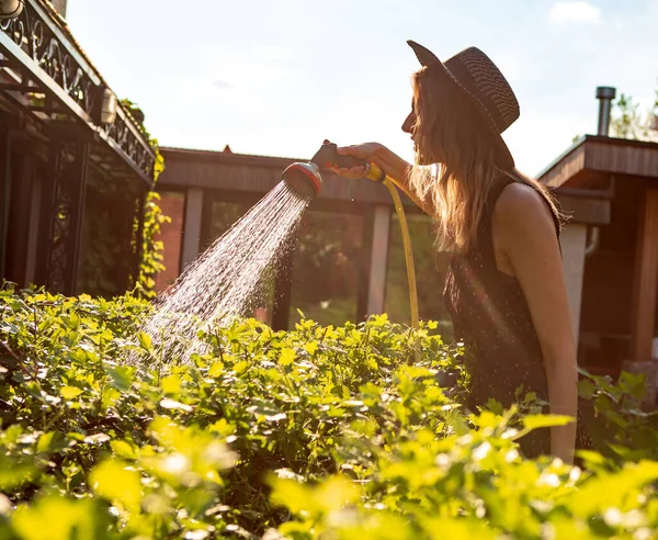 Chica bonita en un sombrero regando plantas con una manguera de jardín en el jardín en verano, fotografía para blog o anuncio —  Fotos de Stock