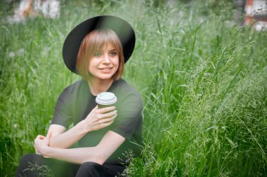 Siyah şapkalı genç kız bahçedeki yeşil çimlerde bir fincan kahveyle, çevre dostu aktif yaşam tarzı