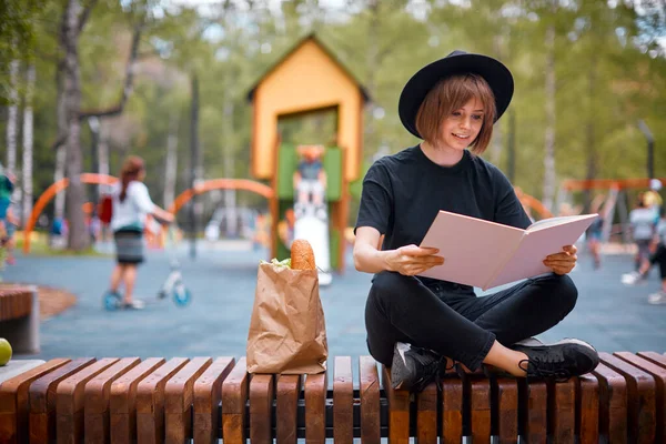 Egy fiatal, aranyos, modern Y-generációs hipszter lány fekete kalapban pihentető könyvet olvas egy városi parkban.. — Stock Fotó