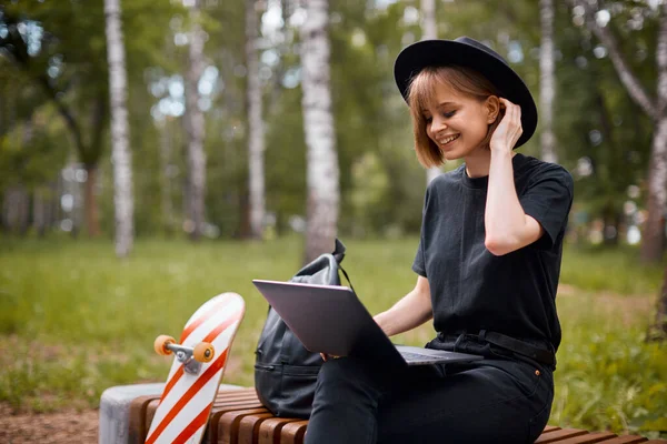 Kreatív nevetés boldog lány blogger munka laptop tartalmat ül padon gördeszka és hátizsák a parkban — Stock Fotó