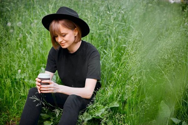 Chica joven y bonita en negro y sombrero con una taza de café o té en la hierba verde, estilo de vida activo ecológico —  Fotos de Stock