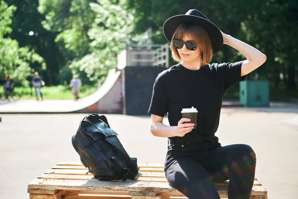 Una joven linda chica hipster milenaria moderna en un sombrero vestido de negro es relajarse en un parque de skate de la ciudad con café para llevar . —  Fotos de Stock