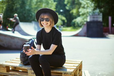 Siyah şapkalı, genç ve modern çağdaş hippi bir kız şehir kaykay parkında kahve içip dinleniyor..
