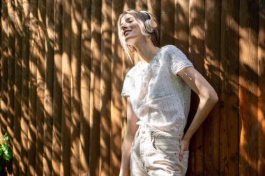 Kablosuz kulaklıklı güzel bir kızın portresi, güneşli bir günde arka bahçede ahşap bir çitin karşısında duruyor..