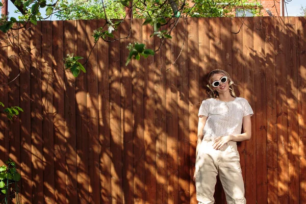 Retrato de una chica bonita con gafas y un pañuelo en la cabeza de pie contra una valla de madera en el patio trasero en un día soleado . — Foto de Stock