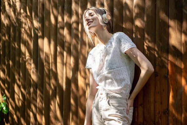 Retrato de una chica bonita con auriculares inalámbricos, de pie contra una valla de madera en el patio trasero en un día soleado . —  Fotos de Stock