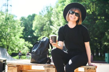 Siyah şapkalı, genç ve modern çağdaş hippi bir kız şehir kaykay parkında kahve içip dinleniyor..