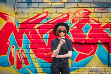 Şapkalı ve güneş gözlüklü güzel kadın renkli grafitiyle tuğla duvarın karşısında duruyor.