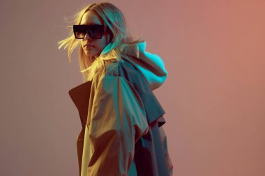 Şık bir genç kız, kapüşonlu, güneş gözlüklü ve pelerinli havalı poz, neon bir arka planda saç çıkıyor..