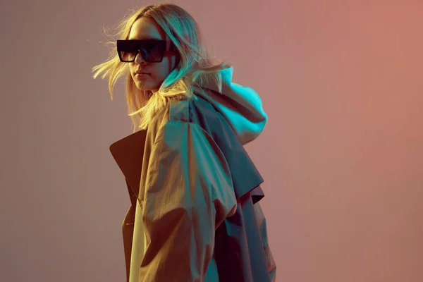 Chica joven con estilo, posando fresco en una sudadera con capucha, gafas de sol y capa con el pelo en desarrollo, sobre un fondo de neón . —  Fotos de Stock