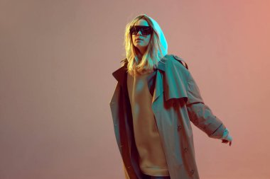 Şık bir genç kız, kapüşonlu, güneş gözlüklü ve pelerinli havalı poz, neon bir arka planda saç çıkıyor..