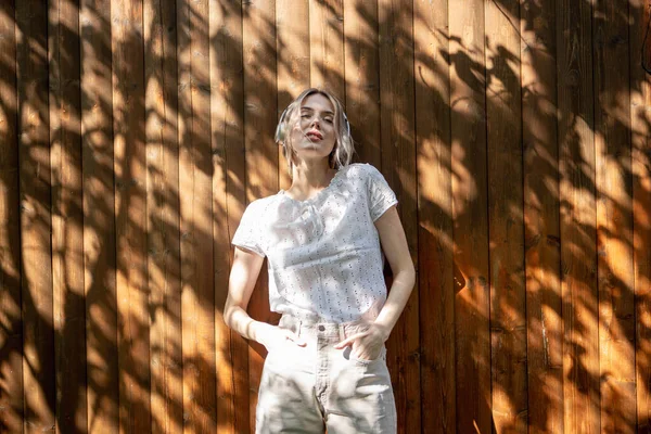 Retrato de una chica bonita con auriculares inalámbricos, de pie contra una valla de madera en el patio trasero en un día soleado . —  Fotos de Stock