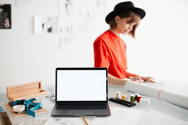 Fiatal lány divattervező kalapban és laptopban, üres képernyővel a stúdióban. Fénykép hirdetési tanfolyamok vagy ruházat — Stock Fotó