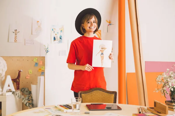 Pretty smiling girl artist in hat stands with drawing in art studio. Photography for ad of art courses or blog