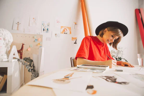 Ung söt flicka konstnär i hatt drar akvareller i studio. Foto för reklam kurser illustrationer eller blogg — Stockfoto