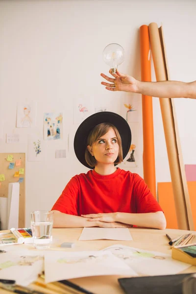 Молодая красивая девушка-художник в шляпе думает и идея приходит в студию. Проницательность символизируется лампочкой в руке — стоковое фото