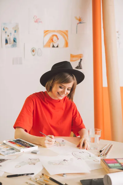 Ung söt flicka konstnär i hatt drar akvareller i studio. Foto för reklam kurser illustrationer eller blogg — Stockfoto