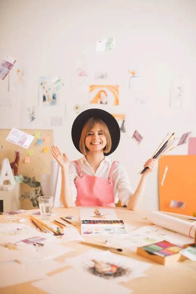 Criativa sorrindo ilustrador menina sentado sob chuva de dinheiro na oficina. Fotografia de conceito sobre arte e freelance — Fotografia de Stock