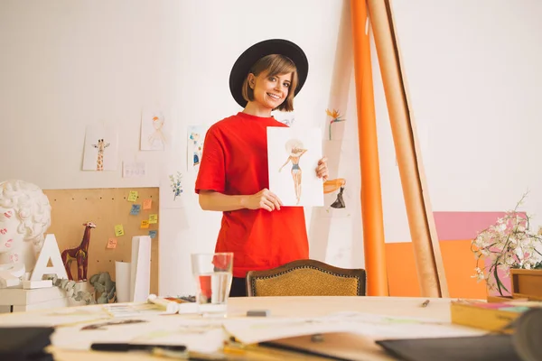 Ganska leende flicka konstnär i hatt stå med akvarell ritning i konst studio. Fotografi för annons av konstkurser eller blogg — Stockfoto