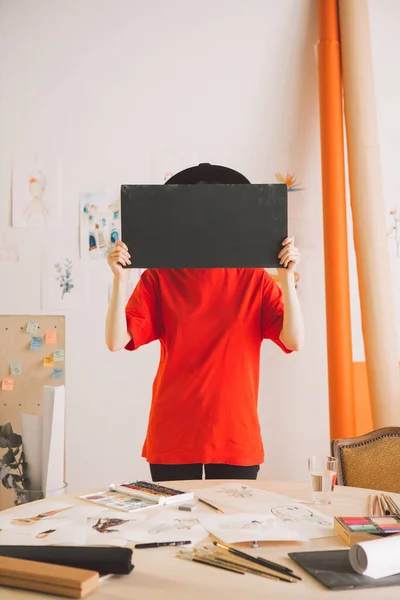 Chica joven en sombrero cubre la cara con cartel de cartón negro con puestos de espacio de copia en el estudio de arte — Foto de Stock