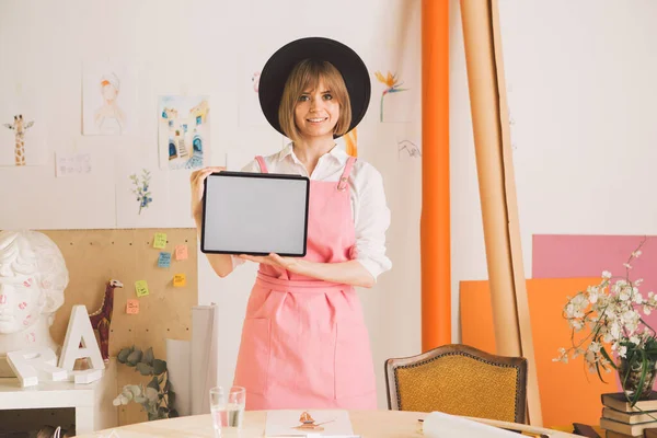 Young smiling pretty girl artist in hat and apron showing blank screen tablet in workshop. Cozy photo for ad or blog