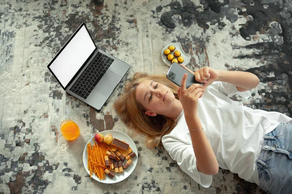 Egy tini lány fekszik a padlón üres laptoppal és telefonnal a közösségi oldalakon. Hangulatos otthoni környezet — Stock Fotó