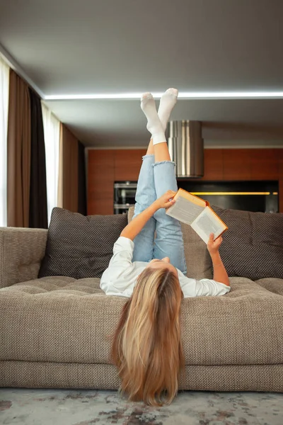 Gündelik giysiler içinde, kanepede ayakları havada uzanmış kitap okuyan tatlı bir genç kız öğrenci. Okula dönelim. Sınav hazırlığı — Stok fotoğraf