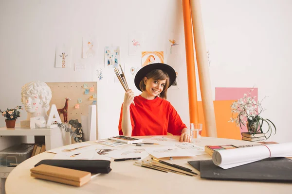 Snygg tjej konstnär i hatt sitter vid bordet med penslar i konststudio. Foto för annonskurser illustrationer eller blogg — Stockfoto