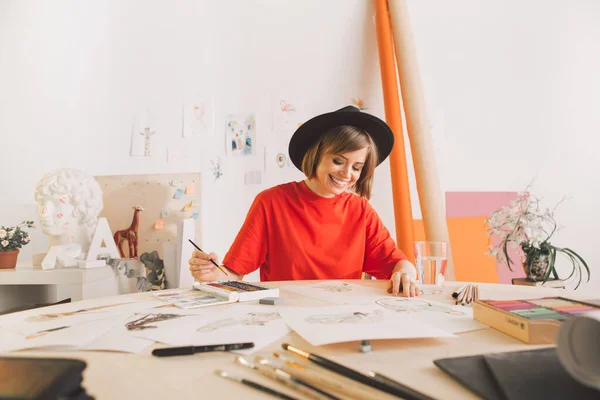 Joven artista chica bonita feliz en el dibujo del sombrero en la mesa en el estudio. Fotografía sobre inspiración y perspicacia — Foto de Stock