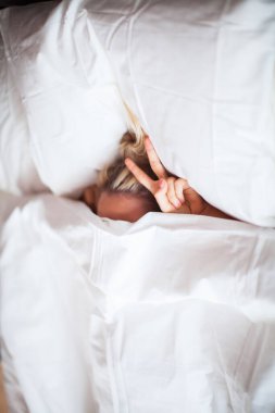 Genç kız beyaz yatakta battaniyenin altında, yatak odasında V işaretli. Sağlıklı uyku, uyku uygulamaları, tembel sabahlar.