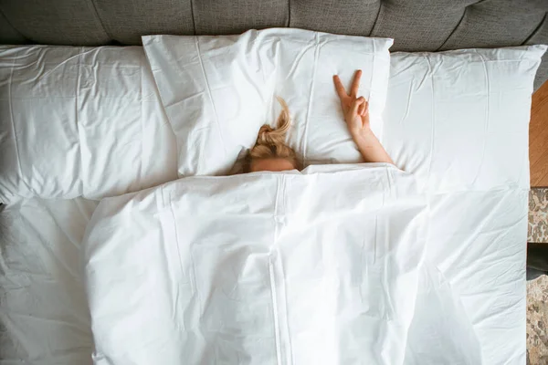 Νεαρή έφηβη κάτω από κουβέρτα σε λευκό κρεβάτι στο υπνοδωμάτιο με ένδειξη χειρονομία V. Υγιής ύπνος, εφαρμογές ύπνου, τεμπέλικο πρωινό — Φωτογραφία Αρχείου