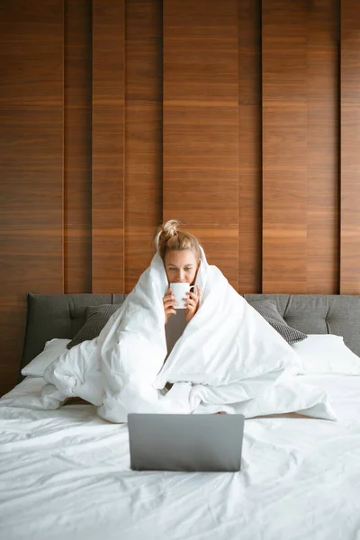 Χαριτωμένο έφηβος κορίτσι κάτω από κουβέρτα σε λευκό κρεβάτι στο υπνοδωμάτιο ξύπνησε και να πίνουν τσάι με φορητό υπολογιστή. Πρωινό στο κρεβάτι, τεμπέλικο πρωινό — Φωτογραφία Αρχείου