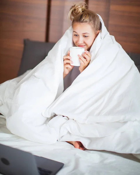 Χαμογελώντας έφηβος κορίτσι κάτω από κουβέρτα στο κρεβάτι στην κρεβατοκάμαρα ξύπνησε και να πίνουν καφέ με φορητό υπολογιστή. Πρωινό στο κρεβάτι, τεμπέλικο πρωινό — Φωτογραφία Αρχείου