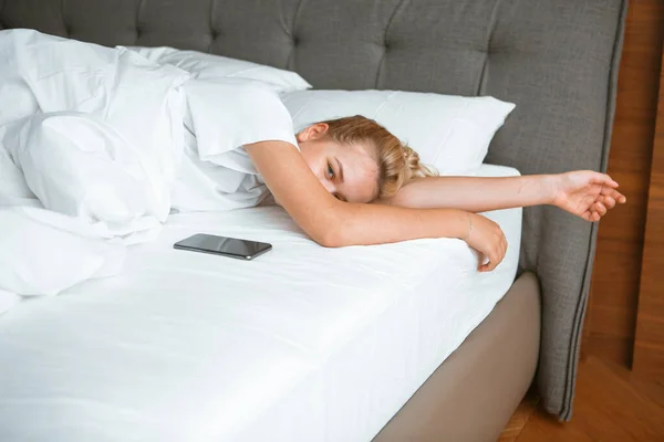 Νεαρή κοπέλα κοιμάται με το τηλέφωνο κάτω από κουβέρτα σε λευκό κρεβάτι στην κρεβατοκάμαρα. Υγιής ύπνος, υγιεινές εφαρμογές ύπνου, τεμπέλικο πρωινό — Φωτογραφία Αρχείου