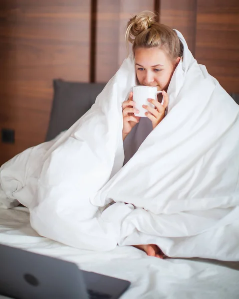 Χαριτωμένο έφηβος κορίτσι κάτω από κουβέρτα σε λευκό κρεβάτι στο υπνοδωμάτιο ξύπνησε και να πίνουν τσάι με φορητό υπολογιστή. Πρωινό στο κρεβάτι, τεμπέλικο πρωινό — Φωτογραφία Αρχείου