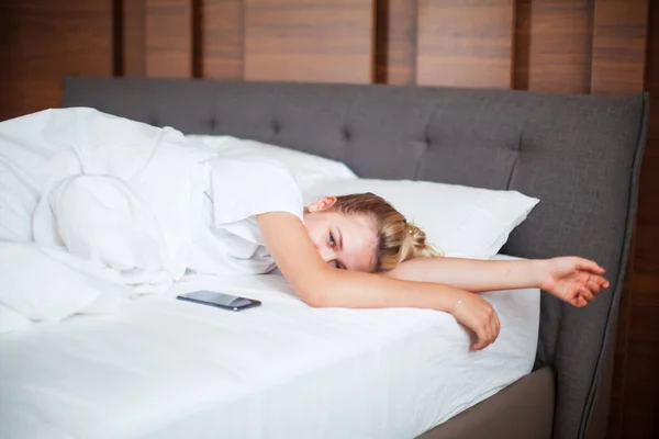 Νεαρό κορίτσι ξυπνάει με το τηλέφωνο κάτω από κουβέρτα σε λευκό κρεβάτι στην κρεβατοκάμαρα. Υγιής ύπνος, υγιεινές εφαρμογές ύπνου, τεμπέλικο πρωινό — Φωτογραφία Αρχείου