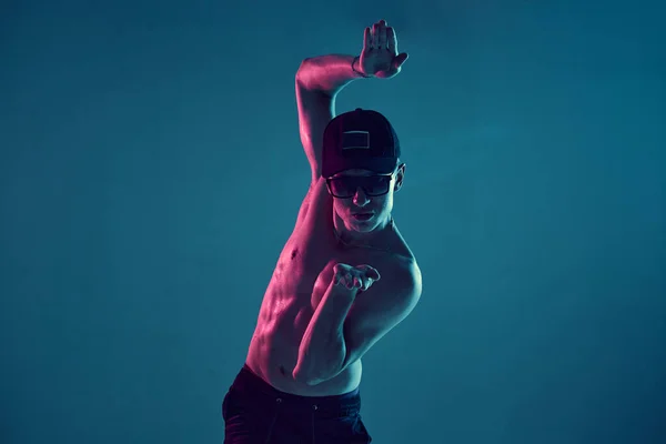 Jeune homme en casquette et lunettes de soleil dansant break dance sans t-shirt en studio en néon bleu. Cours de break dance — Photo