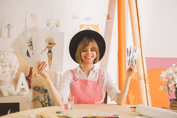 Kreativ glad flicka i hatt artist eller illustratör sitter med pengar och teckningar i sin verkstad — Stockfoto
