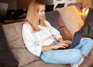 Lotus pozisyonunda kanepede oturan gülümseyen genç kız dizüstü bilgisayarda çalışıyor. Konforlu ev fotoğrafı