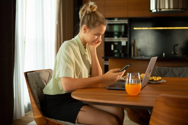 Kreatív aranyos lány blogger laptoppal és okostelefonnal otthon ülve. Hangulatos konyhai környezet — Stock Fotó