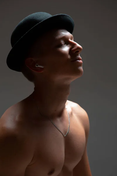 Un joven con sombrero negro y auriculares inalámbricos escucha canciones sin camisa sobre fondo negro. Cartel musical —  Fotos de Stock