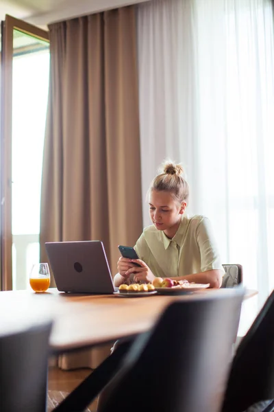 Csinos fiatal modern lány okostelefonnal és laptoppal ül az asztalnál kényelmes otthon — Stock Fotó