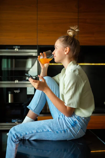 Akıllı telefonlu genç kız sıcacık bir mutfakta meyve suyu içiyor. — Stok fotoğraf
